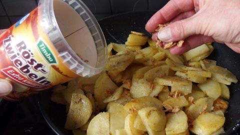 Bratkartoffeln werden in einer Pfanne zubereitet, während Röstzwiebeln darüber gestreut werden.