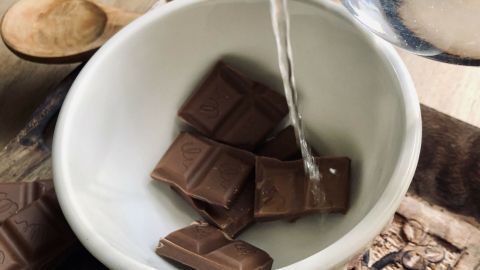 Ein Schüssel mit Schokolade wird mit heißem Wasser übergossen, während ein Holzlöffel daneben liegt.