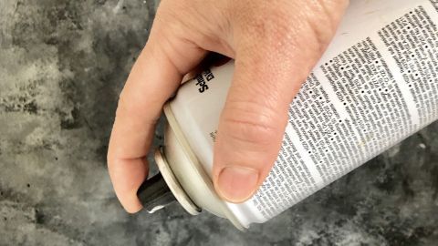 Mit diesem Tipp ist eine angefangene Spraydose auch nach längerer Zeit des unbenutzt seins, wieder parat zum Sprühen. Eine Hand hält eine Spraydose, während sie bereit ist, Farbe auf eine strukturierte graue Fläche zu sprühen.