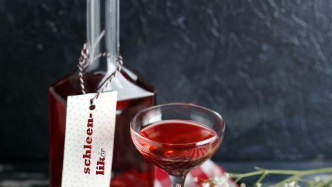 Ein klarer Flasche mit Schlehen-Likör steht neben einem eleganten Glas, umgeben von einer karierten Serviette und Blumen.