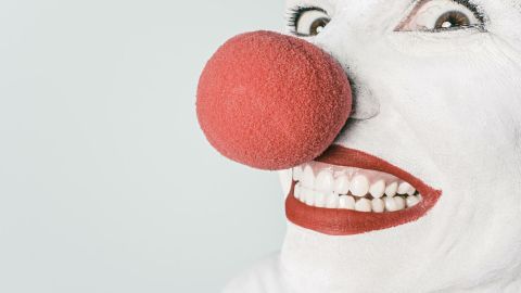 Ein Clown mit roter Nase lächelt breit in die Kamera, während er vor einem neutralen Hintergrund steht.