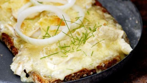 Ein gebackenes Stück Brot mit schmelzendem Camembert, garniert mit Zwiebelringen und frischen Kräutern auf einem Teller.