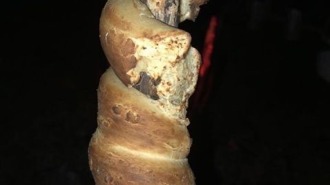 Ein Stockbrot ohne Hefe ist über einem Feuer geröstet. Der Holzstock, umwickelt mit Teig, wird im Dunkeln gehalten.