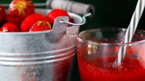 Ein Cocktail aus Erdbeeren steht in einem Glas, während frische Erdbeeren in einem Eimer daneben liegen.