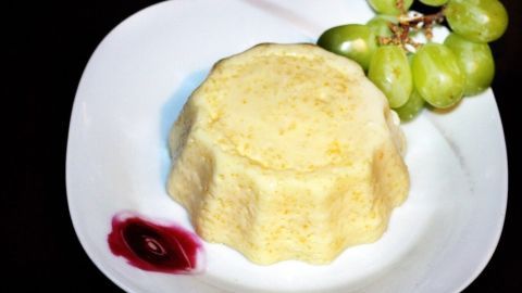 Ein cremiger Grießschaum-Pudding steht auf einem weißen Teller, umgeben von grünen Trauben und einem kleinen Fruchtsoßklecks.
