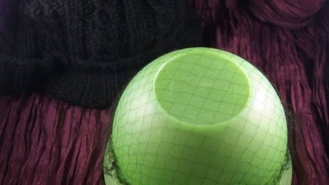 Ein Haarnetz liegt auf einem grünen Objekt, während im Hintergrund eine schwarze Wollmütze und eine lila Textur sichtbar sind.