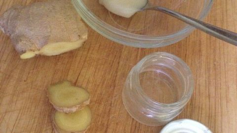 Ein Stück Ingwer liegt neben einer Schale mit cremigem Ingwerhonig auf einem Holzbrett, während ein Löffel bereitsteht.