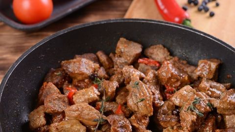 Ein Gulasch aus dem Backofen kocht in einer Pfanne, umgeben von frischen Tomaten und Gewürzen auf einem Holzbrett.