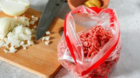 Hackfleisch ist in einem Gefrierbeutel verpackt, neben gehackten Zwiebeln und einem Messer auf einem Holzbrett.