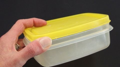 Ein Hand hält einen Kunststoffbehälter mit einem gelben Deckel, der leicht verzogen ist, um ihn wieder in Form zu bringen.