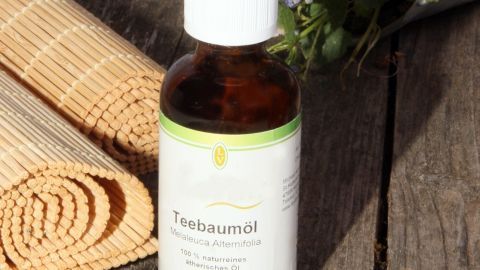 Teebaumöl-Flasche steht auf Holztisch, umgeben von Bambusmatte und Blumen. Text: "Teebaumöl Melaleuca Alternifolia 50 ml."