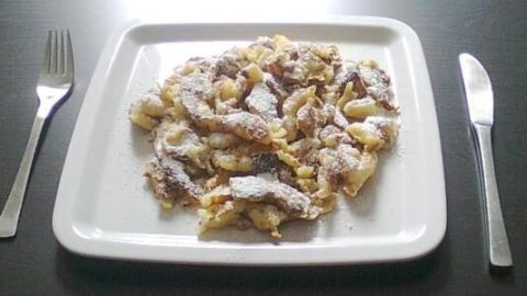 Ein Teller mit Kaiserschmarrn, bestäubt mit Puderzucker, liegt auf einem Tisch. Links und rechts daneben liegen Gabel und Messer.