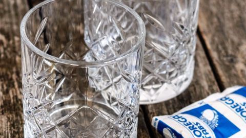 Klar strukturierte Wassergläser stehen auf einem Holzuntergrund, neben ihnen liegen Reinigungstabletten zur Randentfernung.