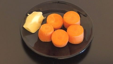 Fünf leuchtend orangefarbene Möhrenstücke liegen auf einem schwarzen Teller, neben einem Stück Fett.