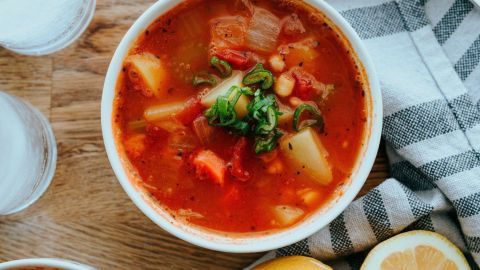 Eine appetitliche italienische Minestrone-Suppe in einer weißen Schüssel wird auf einem Holzbrett serviert, umgeben von Zitronenscheiben und Besteck.