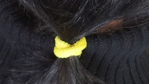 Ein gelbes Haargummi ohne Metallstück hält lange, schwarze Haare zu einem Pferdeschwanz gebunden, vor einem schwarzen Hintergrund.