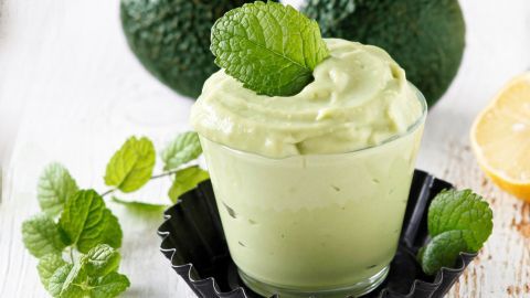 Eine cremige Avocado-Nachspeise steht in einem Glas, umgeben von frischen Minzblättern und einer halben Zitrone.