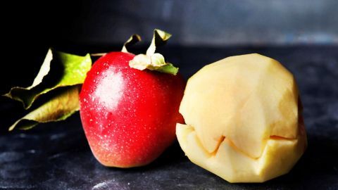 Ein roter Apfel steht neben einem geschälten Apfel, der bereit ist, gegessen zu werden; beide liegen auf einer dunklen Fläche.