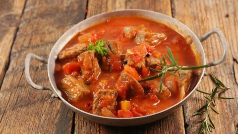 Ein Schaschlikgericht in einer metallenen Pfanne, köchelnd in einer Tomatensauce auf einer rustikalen Holzoberfläche.