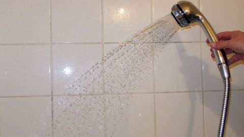 Ein Duschkopf spritzt Wasser auf weiße Fliesen, während eine Hand ihn hält, um die Dusche frisch und rein zu halten.