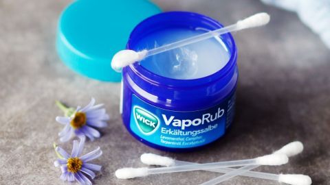 Ein blauer Behälter mit Vick Vaporub steht offen auf einer Tischoberfläche, umgeben von Wattestäbchen und Blumen.