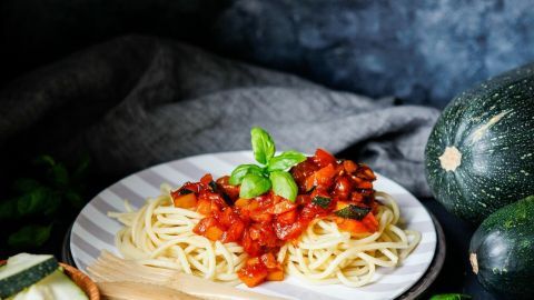 Eine helle Schüssel mit Spaghetti und leckerer Nudelsoße aus Gemüse steht auf einem Tisch, umgeben von Zucchini und frischem Basilikum.