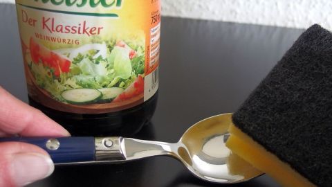 Angelaufenes oder Wasserflecken ganz leicht entfernen - Besteck mit Speiseessig polieren. Eine glänzende Löffel wird mit einem Schwamm gereinigt, während eine Flasche Salatdressing dahinter steht.