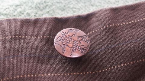 Ein Knopf hält nach dem Annähen länger am Kleidungsstück, wenn man den Faden vorher mit Nagellack bestreicht. Ein runder Knopf liegt auf einem braunen Stoffstück, umgeben von einem hellen Hintergrund, bereit behandelt zu werden.