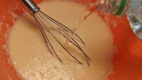 Lockere Pfannkuchen bekommt man, wenn man den Teig mit Mineralwasser zubereitet. Ein Mixer rührt in einer orangen Schüssel einen Teig, während Mineralwasser hinzugefügt wird, um Pfannkuchen zuzubereiten.