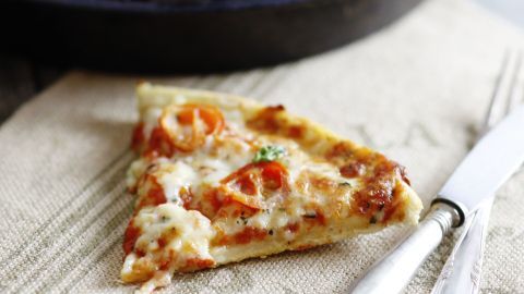 Ein Stück Pizza mit geschmolzenem veganen Käse liegt auf einem Tisch neben einem Besteck auf einem serviettenartigen Tuch.