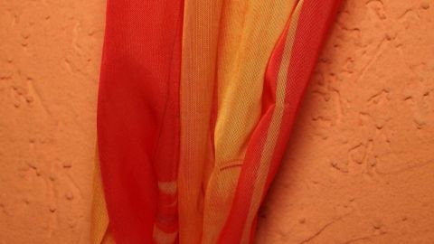 Farbmuster der Wandfarbe zum Aussuchen der Gardinen mitnehmen. Eine bunte Gardine in Rot- und Gelbtönen hängt an einer wandfarbenen Wand und bringt Farbe ins Zimmer.
