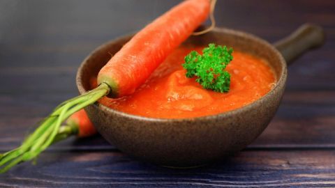 Eine Schüssel mit orangener Karottensuppe, garniert mit frischer Petersilie, steht auf einem Holzuntergrund.