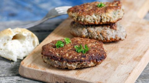 Saftige Frikadellen liegen auf einem Holzbrett mit frischen Kräutern garniert. Im Hintergrund liegt ein Brötchen.