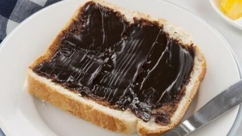 Ein Toast mit dickem, glänzendem Hefeextrakt bestrichen, liegt auf einem Teller neben einem Messer, umgeben von einem karierten Tuch.