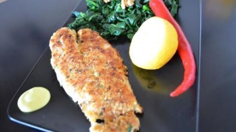 Eine knusprig panierte Fischfilet liegt auf einem schwarzen Teller, umgeben von frischem Gemüse, einer Zitrone und rotem Chili.