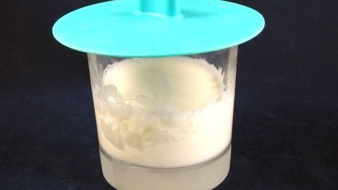 Ein Kunststoffbehälter mit einem türkisfarbenen Deckel enthält warme Milch, die eine cremige Oberfläche zeigt.