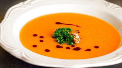 Eine orangefarbene Möhren-Kokos-Suppe befindet sich in einem weißen Teller, garniert mit einem Klecks Kräuter und roten Tropfen.
