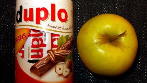 Ein Tipp gegen den inneren Schweinehund - so vermeidet man das Essen von Süßigkeiten. Ein Duppo-Riegel steht neben einem grünen Apfel auf einer schwarzen Oberfläche, die gesunde Wahl gegen den inneren Schweinehund.