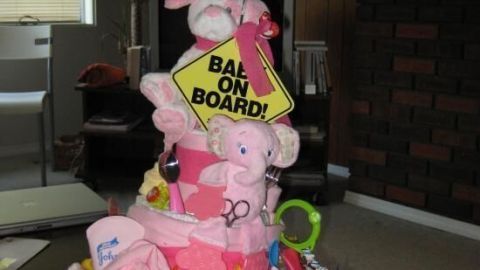 Eine Windeltorte, dekoriert mit Spielzeugen und einem "Baby on Board"-Schild, steht auf einem Tisch in einem gemütlichen Raum.