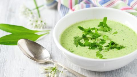 Eine Schüssel Bärlauchsuppe mit frischen Blättern garniert, daneben ein Löffel und weiße Blüten auf einem Holztisch.