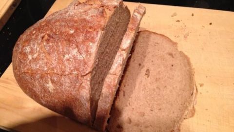 Ein Laib Roggenbrot liegt auf einem Holzbrett mit mehreren geschnittenen Scheiben, umgeben von Krümeln.