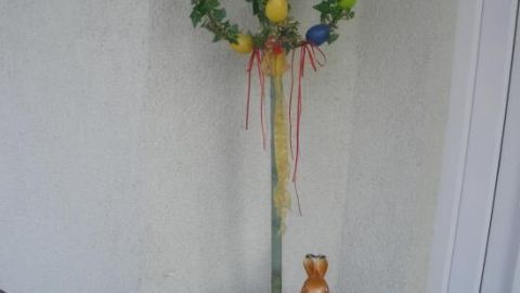 Ein Osterbaum mit bunten Eiern hängt an einem Stock in einem weißen Blumentopf vor einer Wand, umgeben von bunten Blumen.