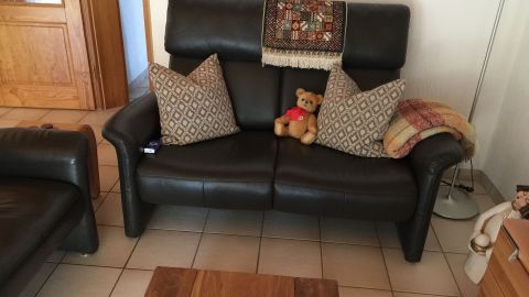 Ein schwarzes Ledersofa steht in einem Wohnzimmer. Darauf liegen Kissen und ein Teddybär. Teppich und Decke ergänzen die Deko.