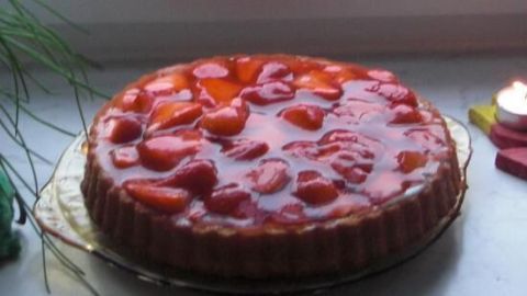 Ein Erdbeertörtchen mit glänzender Oberfläche und frischen Erdbeeren steht auf einer goldenen Tortenplatte vor einem Fenster.