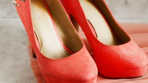 Rote Schuhe stehen auf einem hellen, strukturierten Untergrund neben einem fließenden, rosafarbenen Kleid.