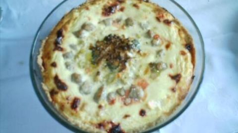 Eine Quiche Rondo in einer klaren Schale, goldbraun gebacken, liegt auf einem weißen Tisch. Sie ist reichhaltig und appetitlich.