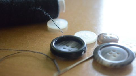 Ein schwarzer Knopf mit vier Knopflöchern liegt auf einem Tisch, während ein Faden und eine Nadel bereit zum Annähen sind.