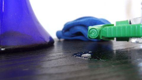 Ein Glasreiniger sprüht auf eine Oberfläche, während ein blauer Lappen daneben liegt, um Staub zu wischen.