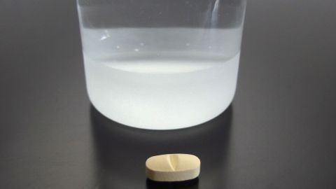 Eine Tablette liegt vor einem halbgefüllten Glas Wasser auf einer schwarzen Oberfläche, bereit zum Schlucken.