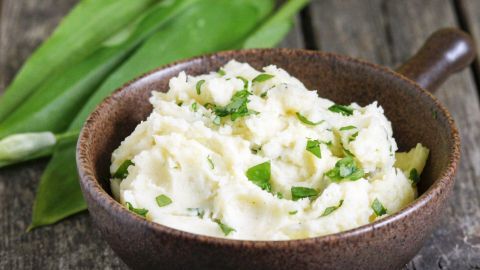 Kartoffelpüree schmeckt mit klein geschnittenem Bärlauch besonders lecker! Cremefarbenes Kartoffelpüree mit frischem Bärlauch, serviert in einer braunen Schüssel auf rustikalem Holztisch.